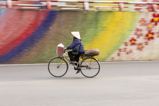 View of a person wearing a traditional conical hat riding a bicycle past a vibrant colorful mural in Hanoi, Ha Noi, Vietnam.