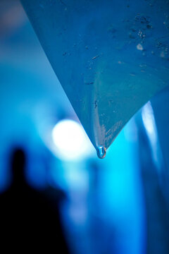 View of the melting ice of the Rhone Glacier with a water droplet forming on a sharp blue ice edge and a blurred silhouette of a person in the background in Obergoms, Valais, Switzerland.