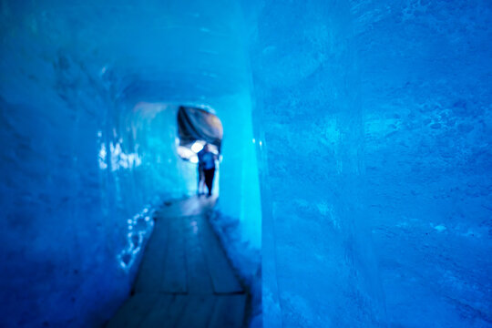 View of the ice tunnel inside the Rhone Glacier with blue glacial walls and a wooden walkway leading to a bright exit in Obergoms, Valais, Switzerland.