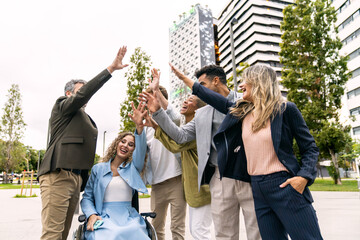 Diverse business people high fiving, celebrating teamwork, success, and inclusion in a city park with office buildings