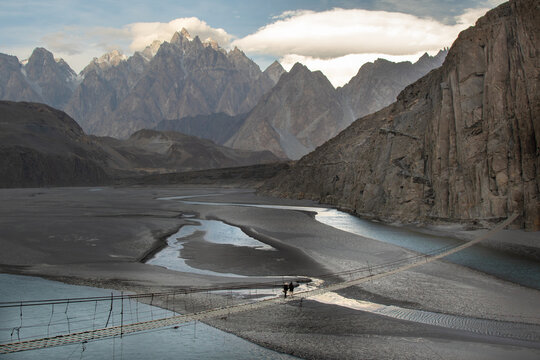 View of the Hussaini Hanging Bridge over the Hunza River with the Passu Cones in the background and two people walking across the bridge in Hunza Nagar, Gilgit, Pakistan.