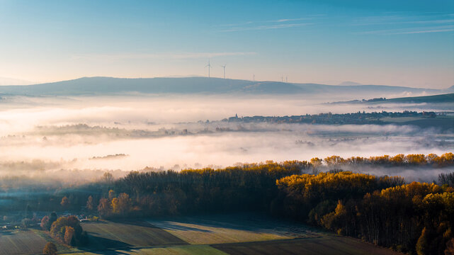 Aerial view of thick morning mist covering a valley with autumn trees and wind turbines on rolling hills under a clear blue sky in Heringen, Hessen, Germany.