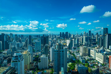 Fototapeta premium Wide aerial view of Bangkok, Thailand, with dense high-rise buildings spread across the city beneath a vivid blue sky and scattered white clouds.