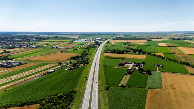 Aerial view of a straight highway stretching through a patchwork of green and golden agricultural fields under a blue sky in Sremska Mitrovica, Serbia.
