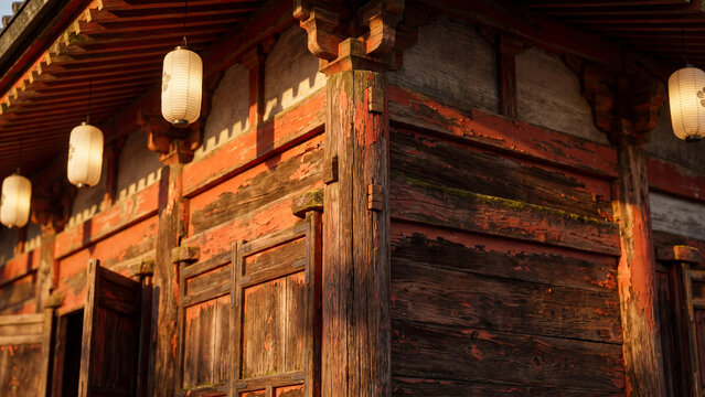 Traditional weathered wooden temple structure featuring worn red paint and glowing white paper lanterns hanging under the eaves during the golden hour sunset light in a peaceful historic Japanese sett