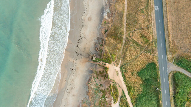 Aerial view of the turquoise waves crashing on the sandy shore of Compton Bay with rugged cliffs and a coastal road in Newport, Isle of Wight, United Kingdom.