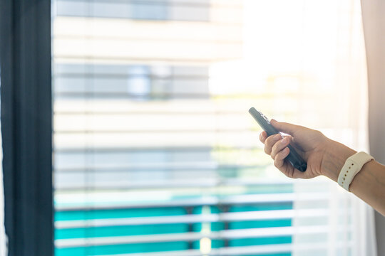 Woman controlling exterior window blinds with a remote control inside a modern apartment. Smart home automation and indoor climate control concept.