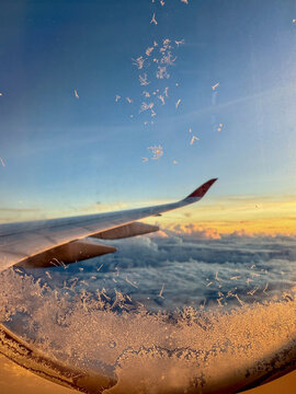 Aerial view of an airplane wing and ice crystals on a window above a thick layer of clouds during sunset in Africa.