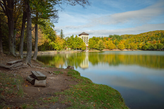 View of Pocuvadlo Lake surrounded by vibrant autumn trees and a hotel in the background with wooden benches on the shore in Banska Stiavnica, Banskobystricky kraj, Slovakia.