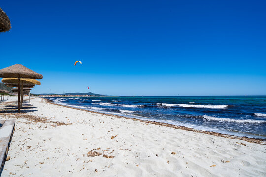 View of Spiaggia Sa Petra Ruja with straw umbrellas on white sand, turquoise sea waves, and kitesurfers in the distance under a clear blue sky in Siniscola, Sardegna, Italy.