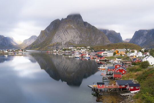 View of the traditional red rorbuer cabins of Lofoten reflected in the calm waters of the fjord, with dramatic mountains rising in the background in Nordland, Norway.