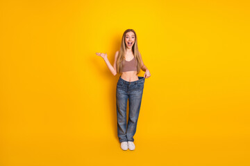 Fototapeta premium Smiling young woman in oversized jeans on yellow background, showcasing successful weight loss and casual style