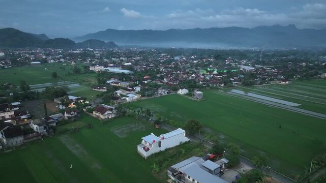 Aerial view of the town of Pacitan surrounded by lush green rice fields and distant mountains under a soft twilight sky in Pacitan, East Java, Indonesia.