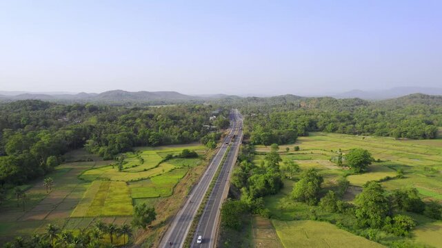 Daylight outdoor aerial drone movement shot of a national highway with vehicles passing through small Indian village surrounded by lush green forest, trees, and farmland in the Konkan region in season
