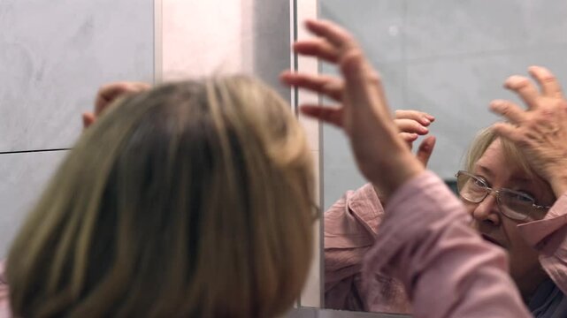 Mature woman looking in a bathroom mirror and checking her hair with her hand. Hair care, self-care and aging beauty concept.