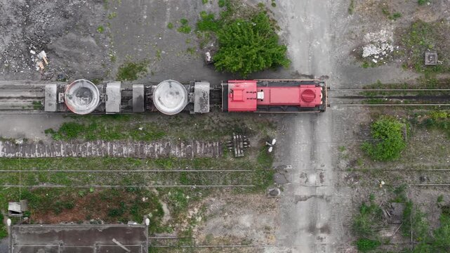 An aerial view captures a red industrial train with large cargo wagons on parallel railway tracks, suggesting heavy transport and industry.