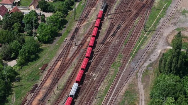 An aerial view captures a vibrant red freight train moving along railway tracks through a rural junction in Hungary, Europe.