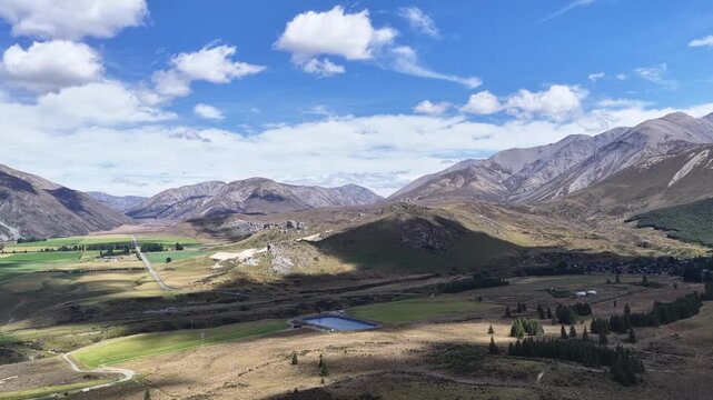 Aerial drone footage over a wide alpine valley in New Zealand with green farmland tussock grasslands rocky mountains and snow capped peaks under a blue sky.