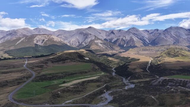 Aerial drone footage over an arid alpine valley in New Zealand with golden tussock grasslands rocky mountains a winding road and river under a dramatic blue sky.