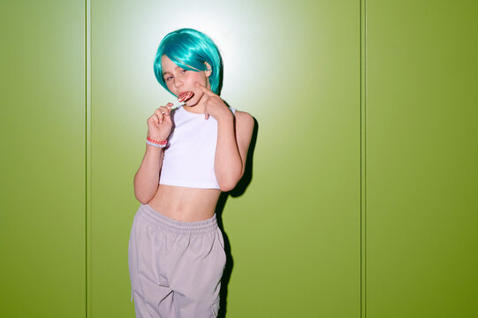 Young girl with vibrant turquoise wig and white crop top playfully licking lollipop against a bright