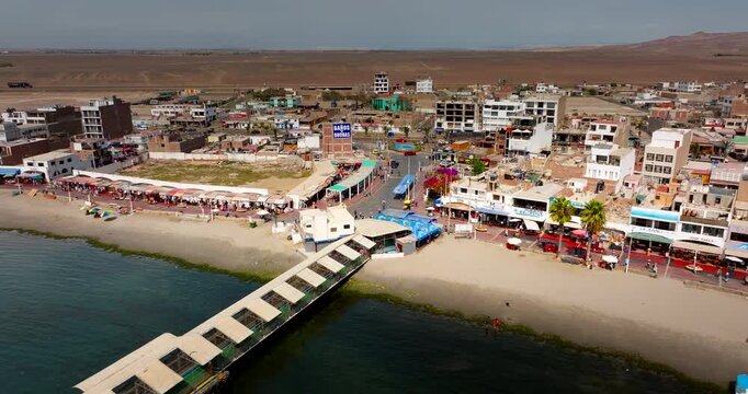 Long pier extending into ocean water near coastal town buildings and beach in Paracas, Peru. Aerial drone