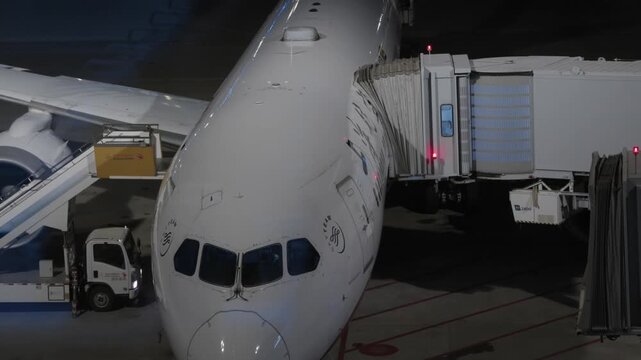 Airplane nose docked at jet bridge with ground vehicles at airport terminal at night