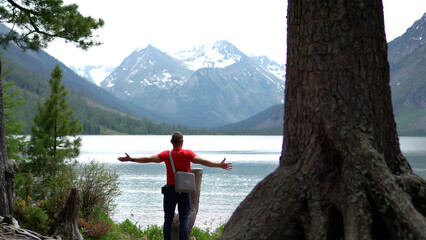 Obraz premium A male adventurer in a red t-shirt standing with outspread arms on an alpine lake shore, celebrating freedom against a majestic snow-capped mountain backdrop.