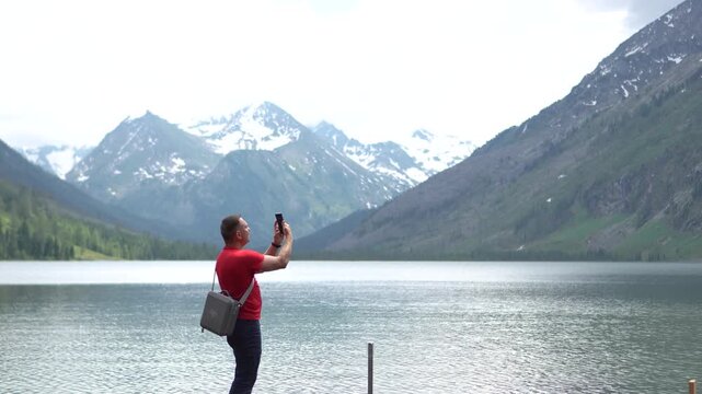 A panoramic medium shot of a male hiker in a red t-shirt spinning in place while using his smartphone to film a 360-degree video of majestic Altai mountains.