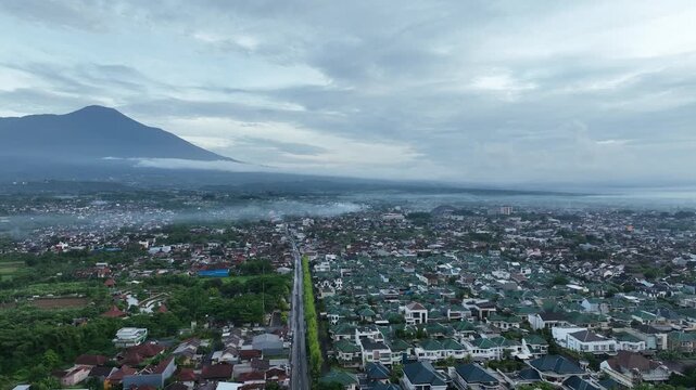 Aerial view of the urban landscape of Purwokerto with Mount Slamet in the background, featuring residential rooftops and a main road in Purwokerto, Central Java, Indonesia.