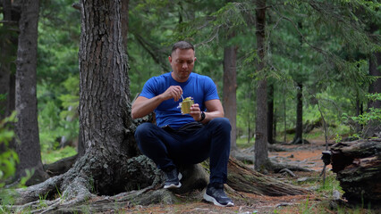 Fototapeta premium A focused male hiker in a blue t-shirt sitting on cedar roots and opening a tin can of food in the Altai forest.
