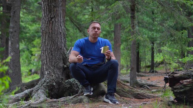A focused male hiker in a blue t-shirt sitting on cedar roots and opening a tin can of food in the Altai forest.
