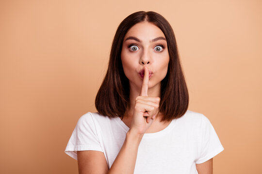 Shh gesture portrait of a young woman in a white tshirt with a beige background expressing secrecy curiosity and playfulness