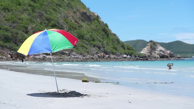 Beach umbrella, colorful parasol on white sand beside turquoise sea along tropical coastline, summer vacation travel scene, gentle waves, mountain island backdrop, clear blue sky