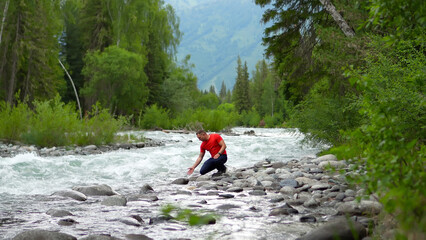 A man travels through the Altai Mountains. A man in red stands on the bank of a mighty mountain river, touching the water, washing himself, and refreshing himself after a long journey. The man enjoys  © OlTarakanov