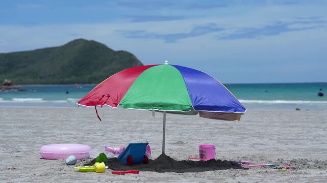 Beach travel umbrella on sand ocean coastline, summer vacation scene with colorful parasol shade, bucket shovel sand toy, inflatable ring, calm water, blue sky, mountain island horizon