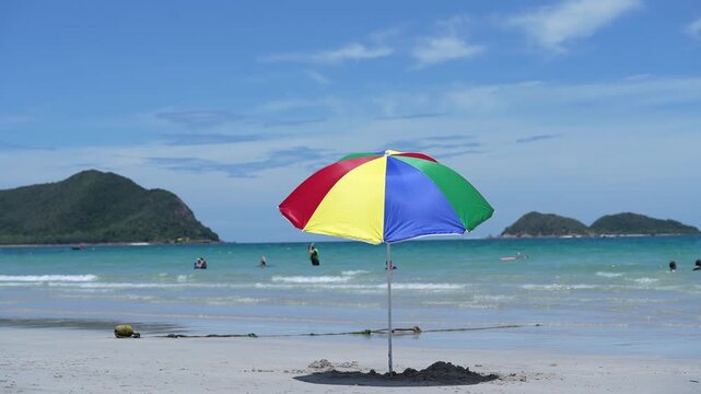 Beach umbrella and parasol on tropical shoreline, soft sand beside ocean wave, island hills, blue sky travel destination, calm seascape with swimmers, bright summer vacation relaxation