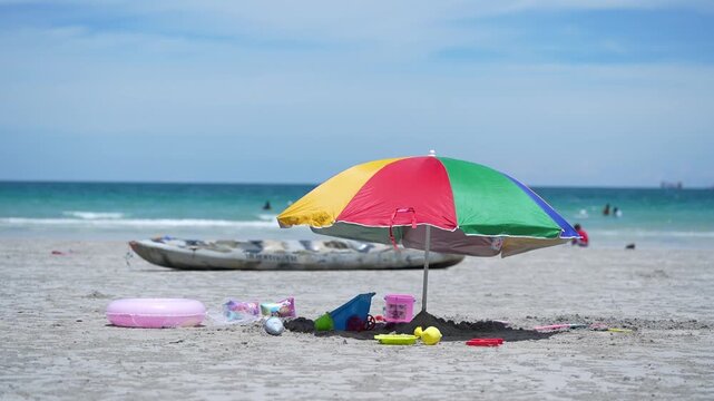 Beach umbrella on sand by ocean and sky, kayak and toy set for vacation, colorful sunshade on quiet shoreline with bucket shovel, inflatable ring, paddle boat, summer travel scene