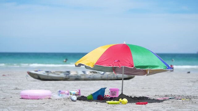 Beach umbrella sand ocean toy kayak summer vacation, colorful parasol on shoreline, sandcastle bucket shovel inflatable ring on sandy beach, turquoise sea gentle waves blue sky copy space