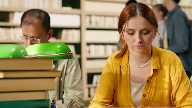 Worried young woman studying materials on laptop at library table, between tall stacks at library, Student showing academic stress and exam preparation workload pressure, supporting study session.