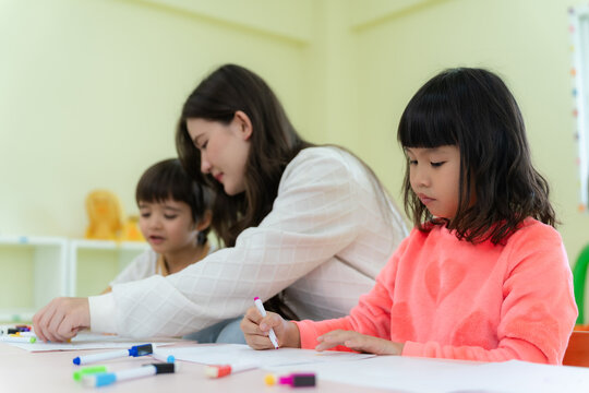 Focused little Asian girl drawing with markers during an art lesson in kindergarten classroom.