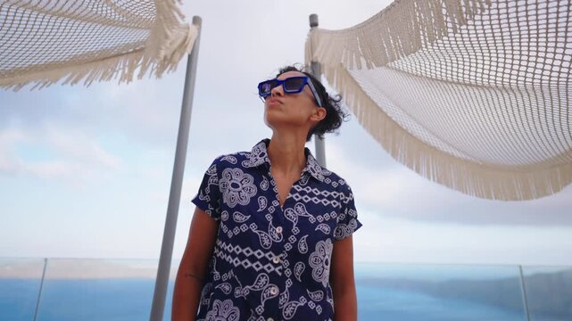 Stylish Indian woman in blue geometric sunglasses and navy paisley print shirt stands confidently between woven fringed parasols at luxury seaside resort with glass balcony and calm ocean backdrop.