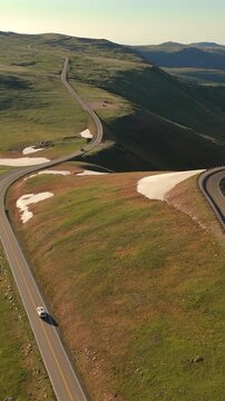 Vertical Screen: Beartooth Highway winding through alpine meadows with melting snow patches in Montana USA. Cars travel along scenic mountain road with golden hour light.