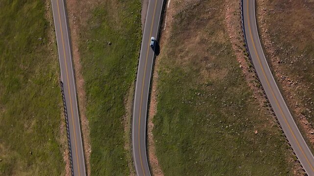 Aerial of Beartooth Highway Montana USA shows white car driving on winding mountain road. Drone footage captures parallel roads with yellow center lines and snow patches on alpine terrain.