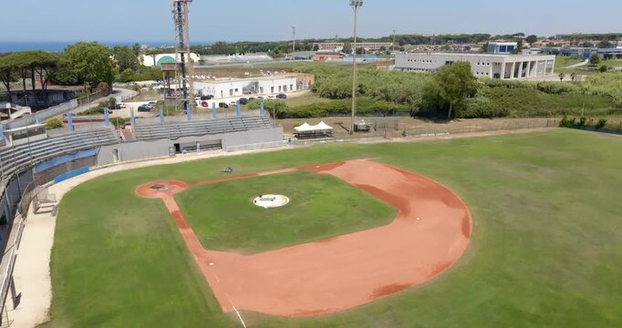 Aerial drone view of an empty baseball stadium on a sunny day, showcasing the distinctive red clay diamond, green grass outfield, concrete grandstands, pitcher's mound, and clear baseline markings.