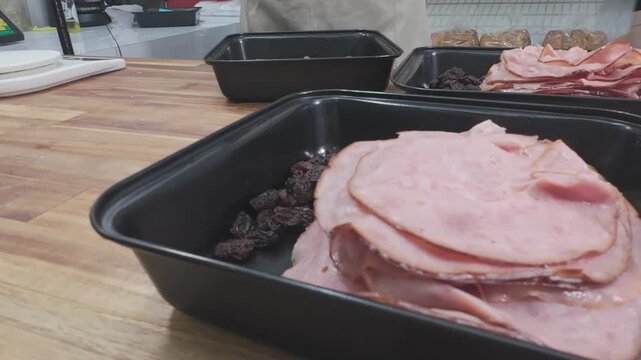 systematic view of black trays filled with sliced ham, raisins, and olives, prepared for the assembly of traditional handmade artisan bread loaves.