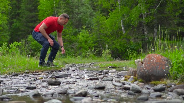 A man is traveling through the Altai Mountains. A man in red is refreshing himself on the bank of a mountain-forest river, washing his face, and throwing a stone into the current.