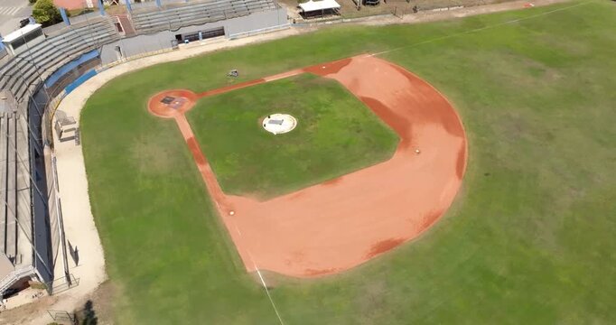 Aerial view of a baseball diamond from the home plate area, highlighting the white pitcher's mound, red clay infield, and concrete stadium grandstands under a bright summer sky. There's nobody.