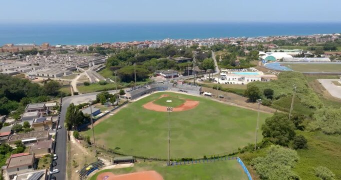Aerial panorama of a beautiful baseball field nestled within a coastal sports complex, showcasing the green outfield, red dirt infield, and a stunning blue sea horizon. It is a sunny summer morning.
