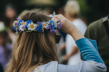 Naklejka premium Young woman with long brown hair wearing a floral crown made of daisies and wildflowers adjusts her hair while standing outdoors in a vibrant festival setting