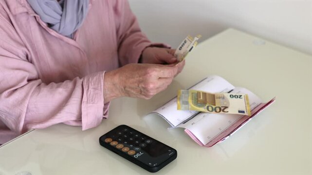 Senior woman counting euro banknotes at home with bills and calculator on smartphone. Close-up of household budgeting, retirement finances, pension money management and cost of living concept.